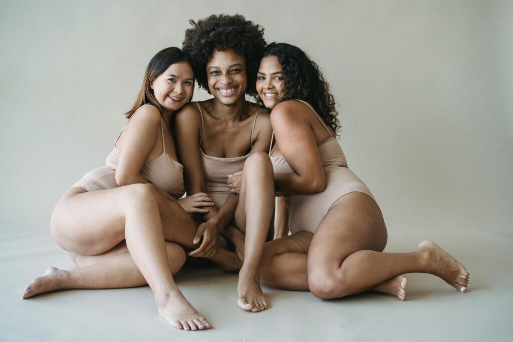 Three women embracing diversity and body positivity in neutral bodysuits on a studio set.