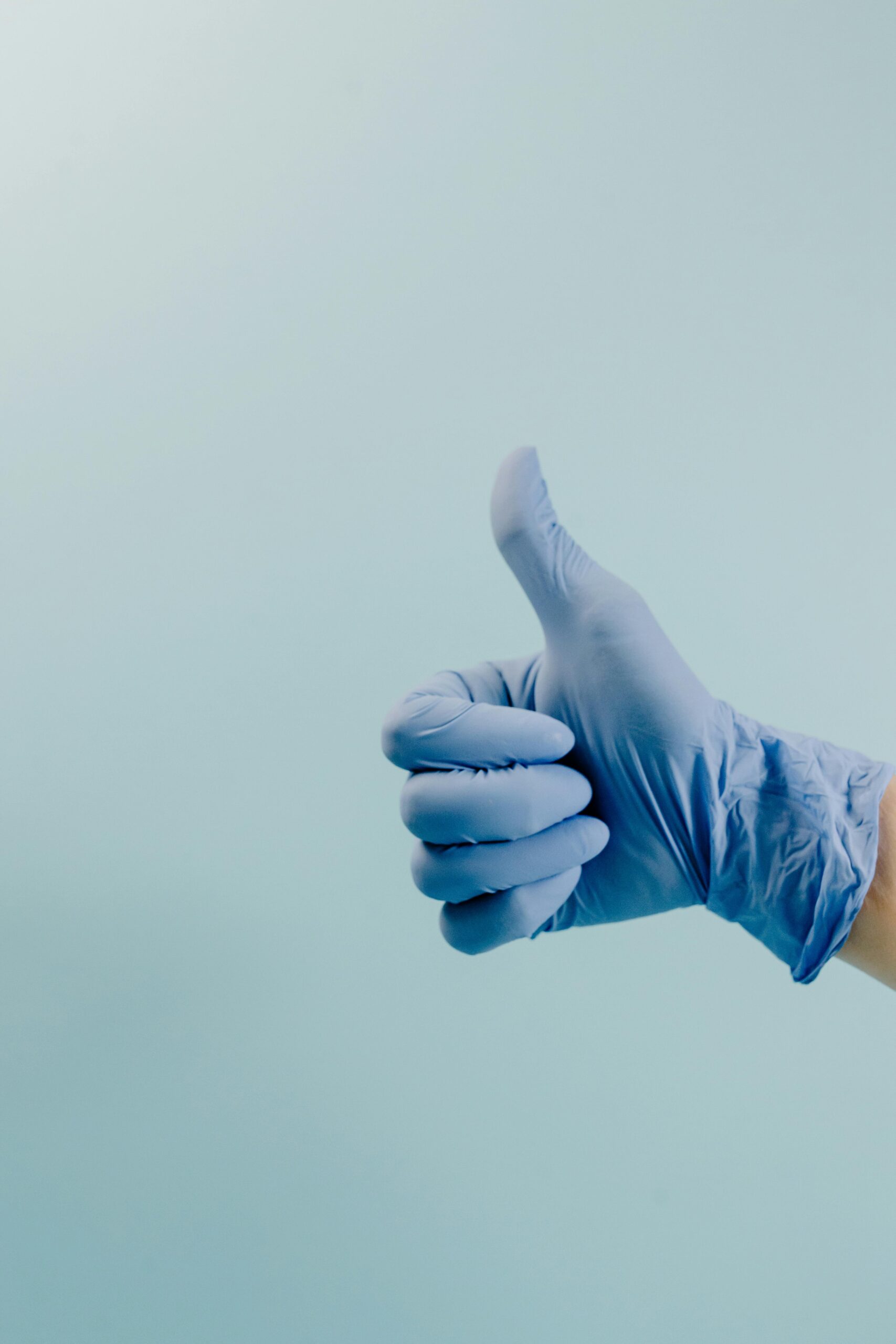 A blue surgical gloved hand showing a thumbs up gesture against a cyan background.